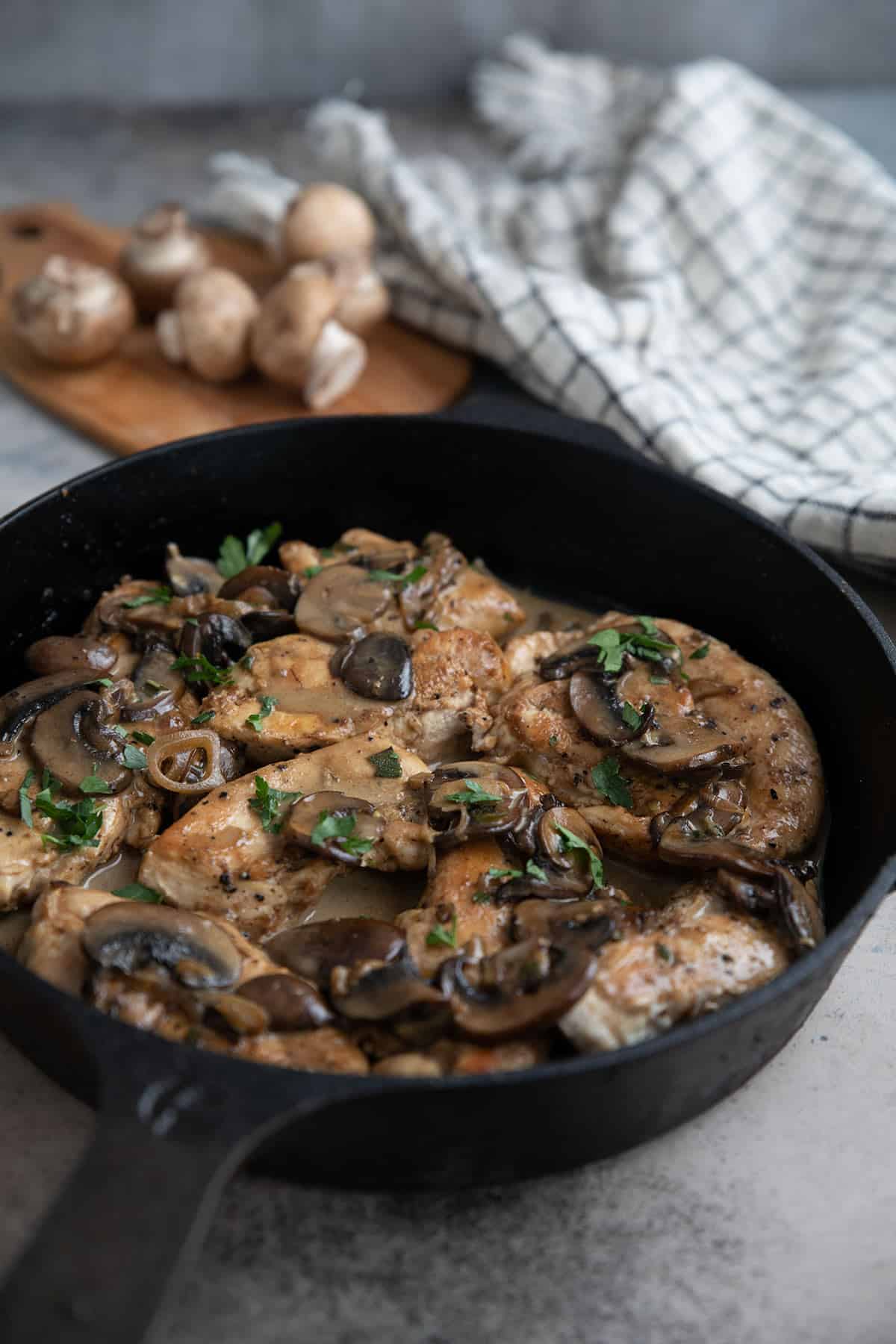 A pan of Keto Chicken Marsala sits on a concrete table with a black and white napkin and mushrooms in the background.