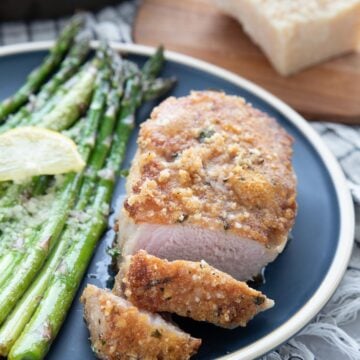 A parmesan crusted pork chop on a blue plate, with two pieces sliced off and some asparagus on the side.