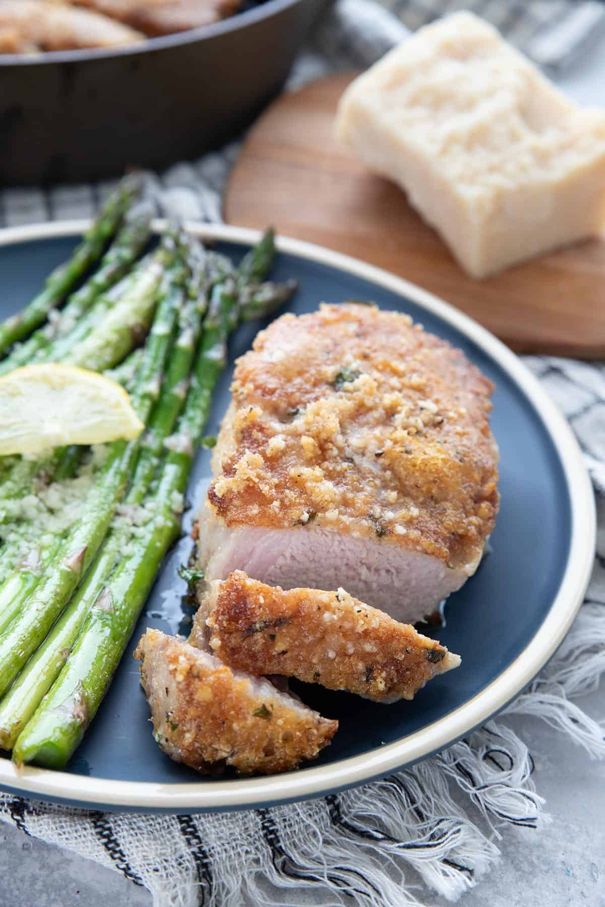 A parmesan crusted pork chop on a blue plate, with two pieces sliced off and some asparagus on the side.