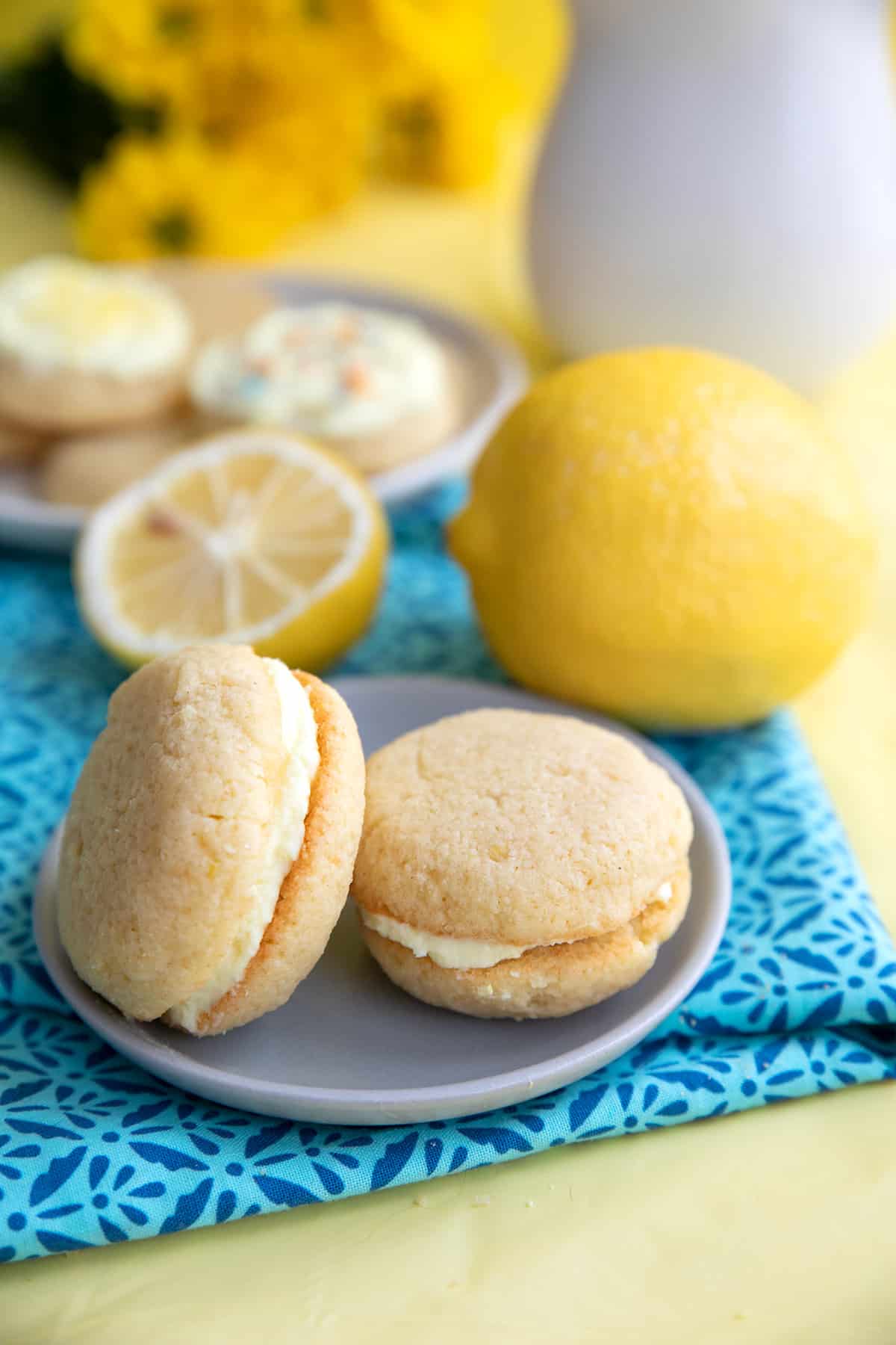 Two keto lemon cookies on a small plate over a patterned blue napkin, with lemons in the background.
