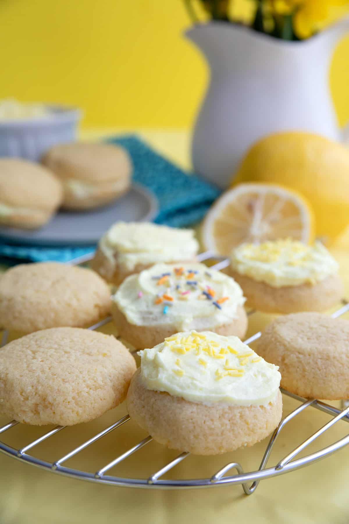 Frosted Lemon Keto Cookies on a wire rack in front of lemons and a vase of yellow flowers.