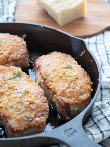 Parmesan Crusted Pork Chops in a cast iron skillet with a chunk of parmesan cheese in the background.