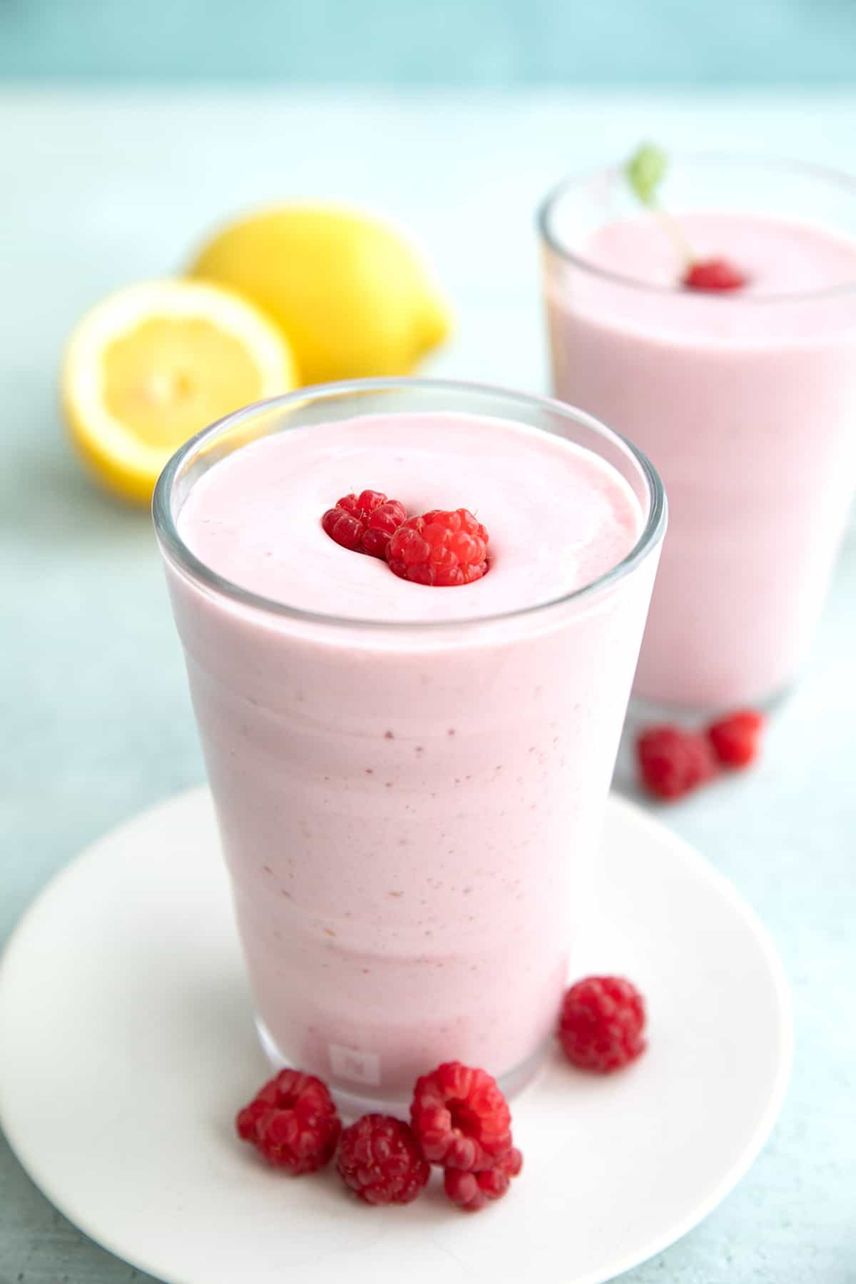 A keto protein shake in a glass on a white plate with raspberries around it and lemon in the background.