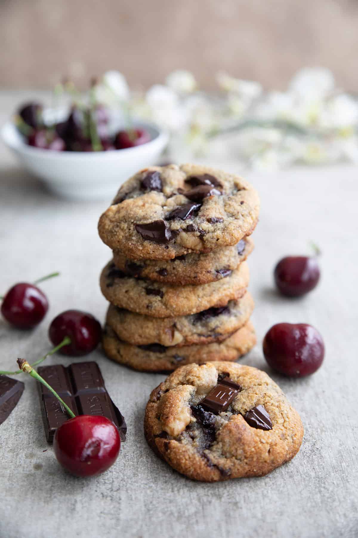 A stack of Keto Cherry Chocolate Chunk Cookies on a concrete table with cherries strewn around.