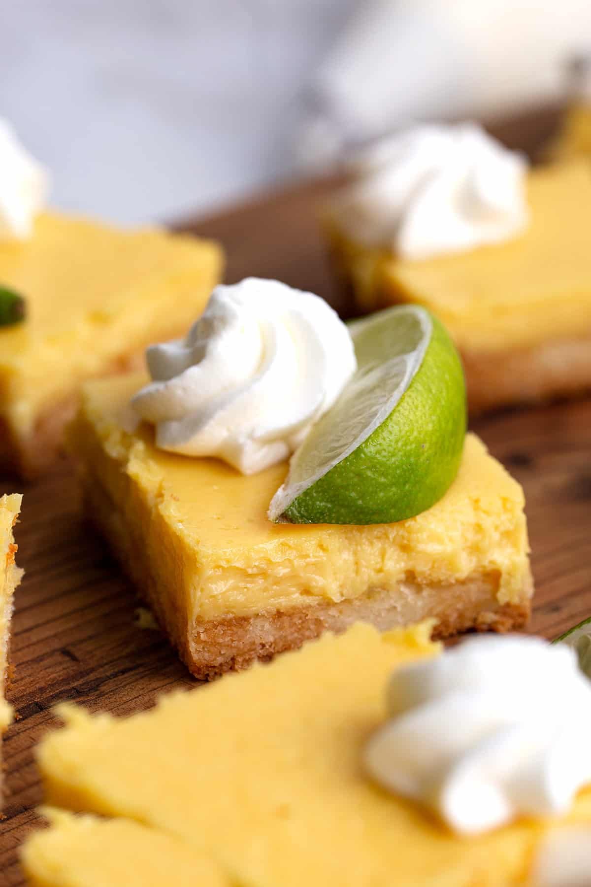 Keto Key Lime Pie bars on a wooden cutting board with whipped cream and lime slices on top.