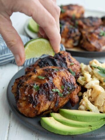 A hand squeezing a wedge of lime over a plate with chipotle lime chicken, avocado slices and cauliflower salad