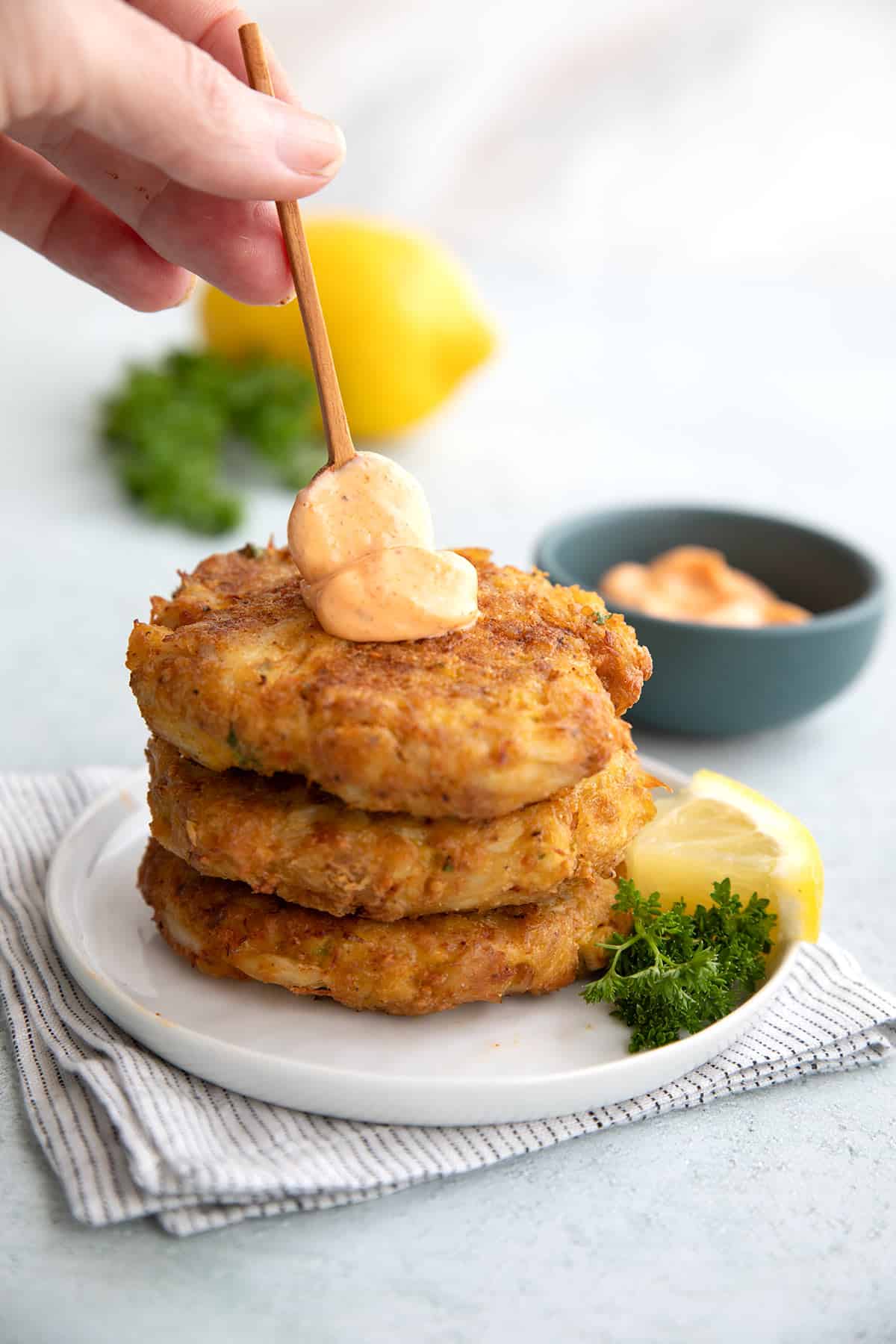A hand spooning spicy mayonnaise on a stack of air fryer crab cakes.