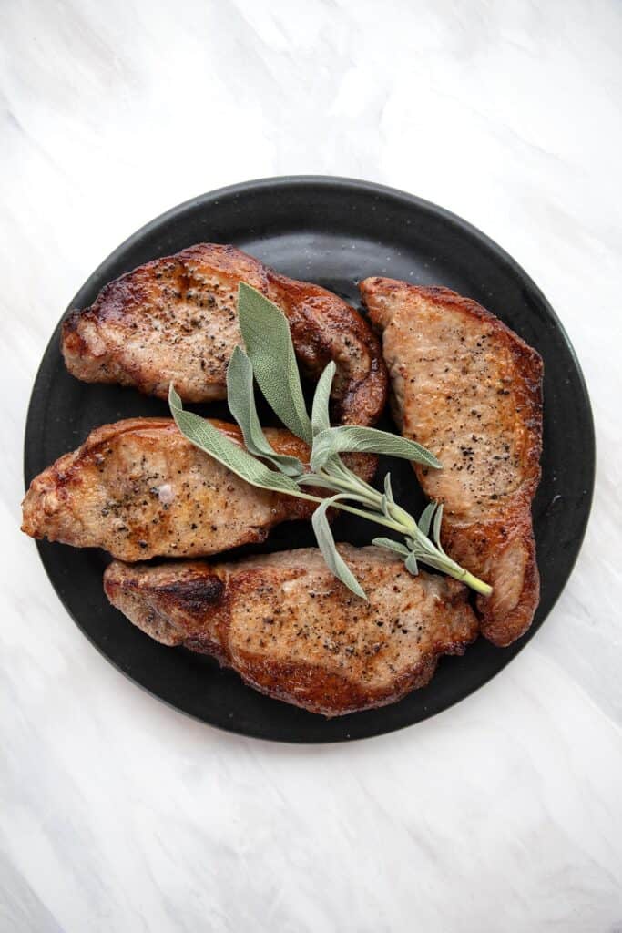 Top down image of four air fryer pork chops on a black plate, topped with a sprig of sage.