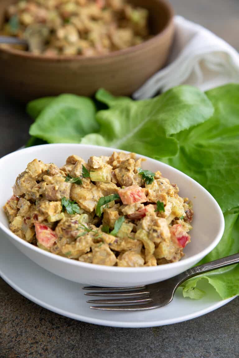 Curried chicken salad in a white bowl, surrounded by lettuce leaves.