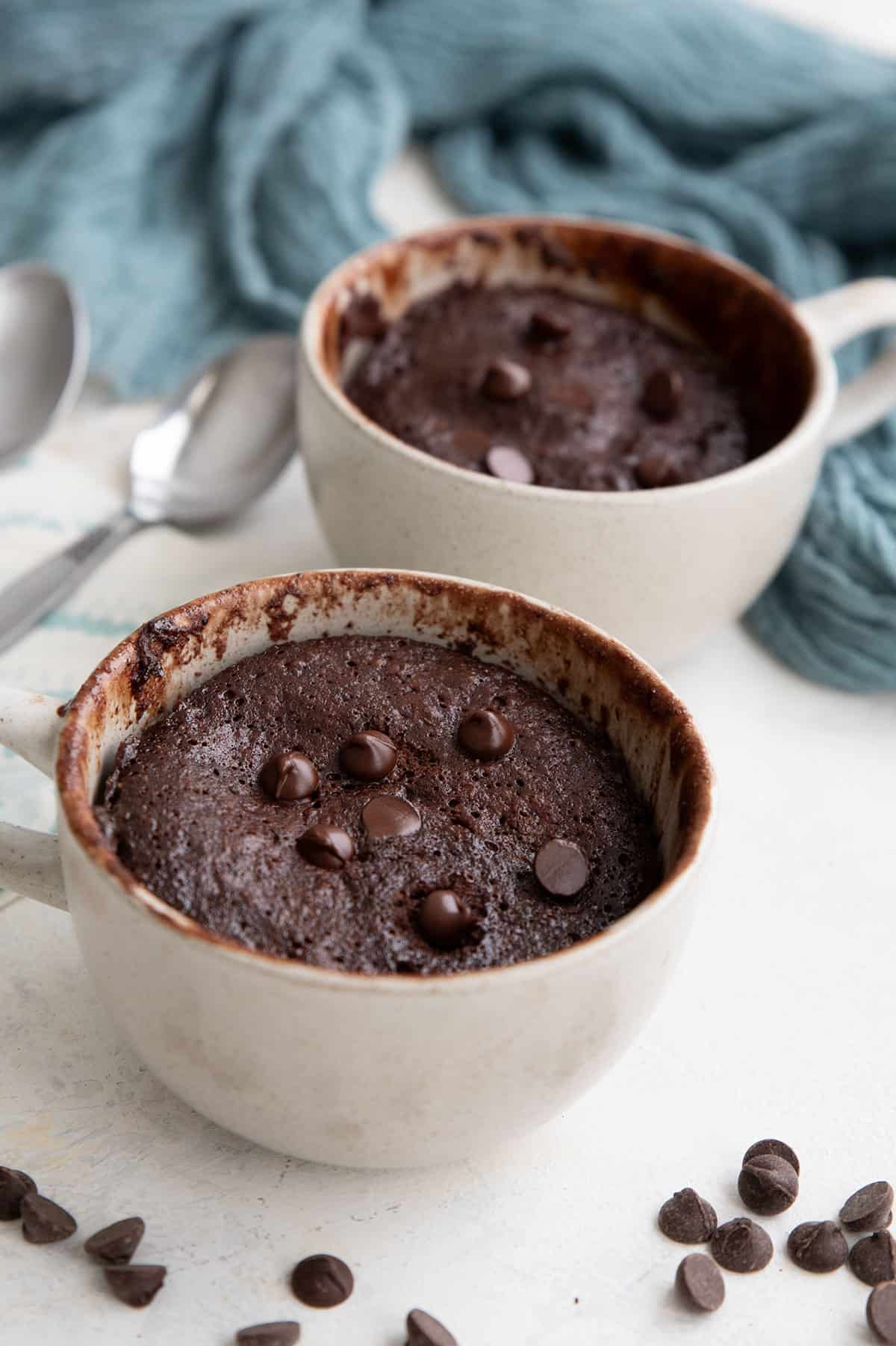 Two protein mug cakes in coffee mugs on a white table with chocolate chips strewn around.