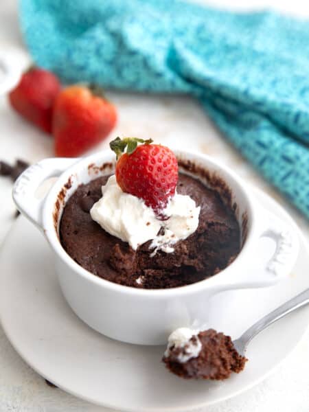 Keto chocolate protein mug cake in a white ramekin with several spoonfuls taken out of it. There is whipped cream and a fresh strawberry on top.