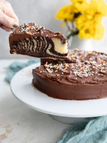 A hand lifting a slice of keto marble cake with chocolate frosting away from the rest of the cake.