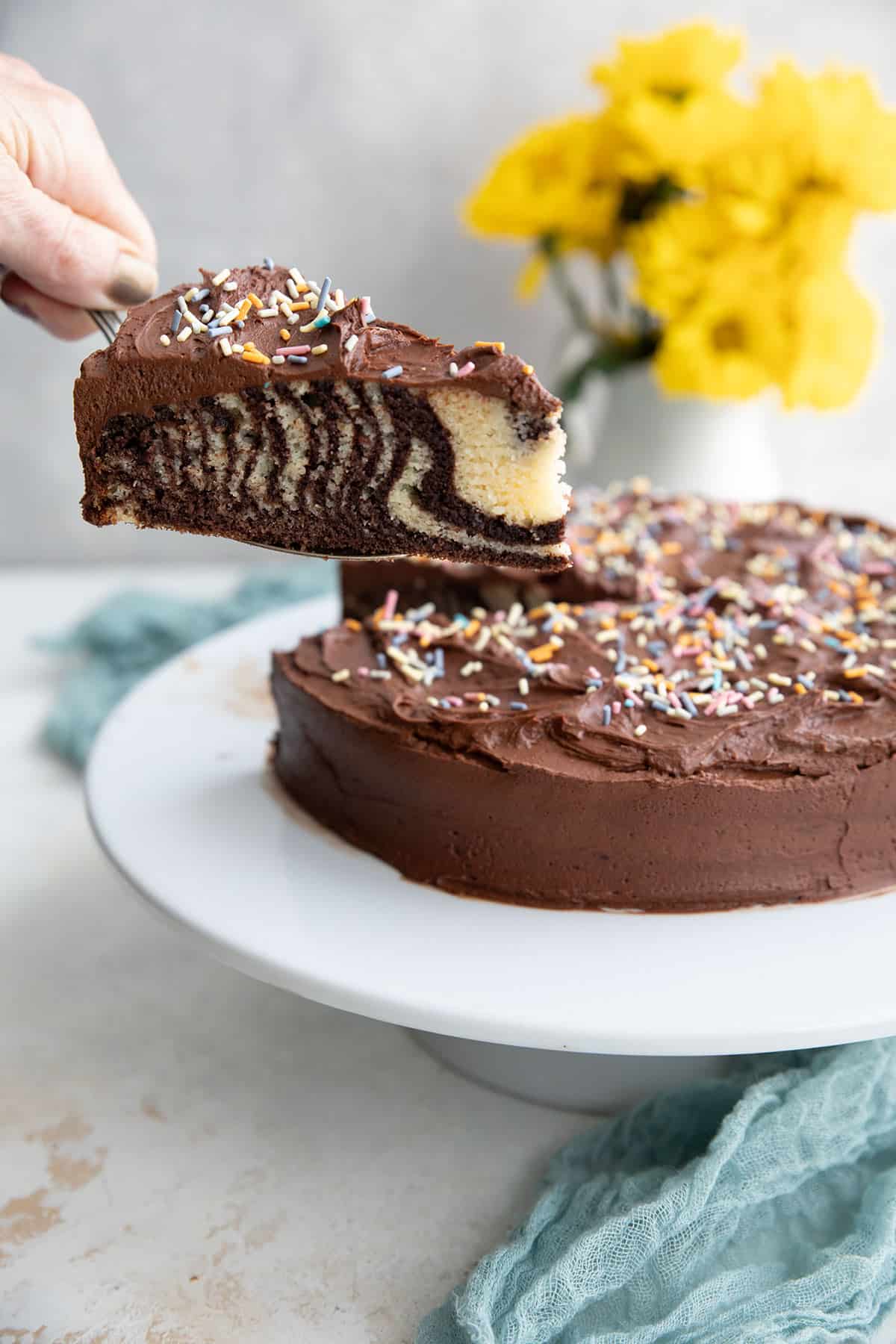 A hand lifting a slice of keto marble cake with chocolate frosting away from the rest of the cake.