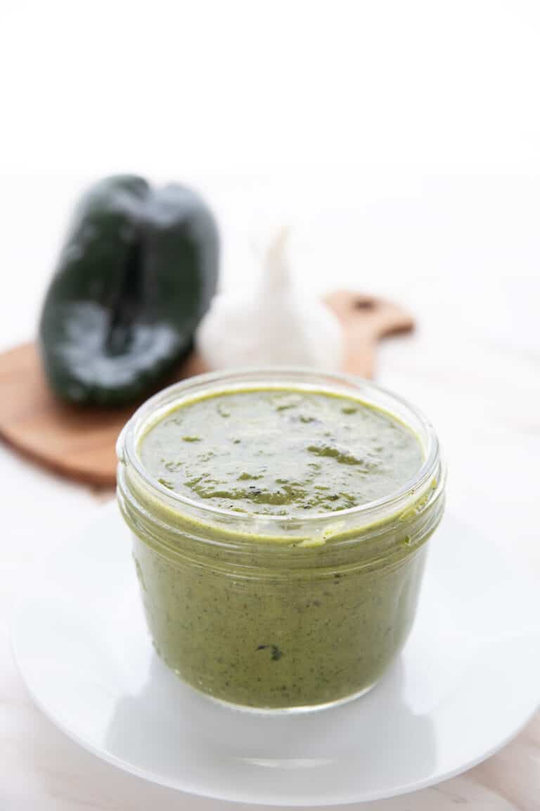 A jar of Poblano Sauce on a white plate in front of a poblano pepper and a head of garlic.