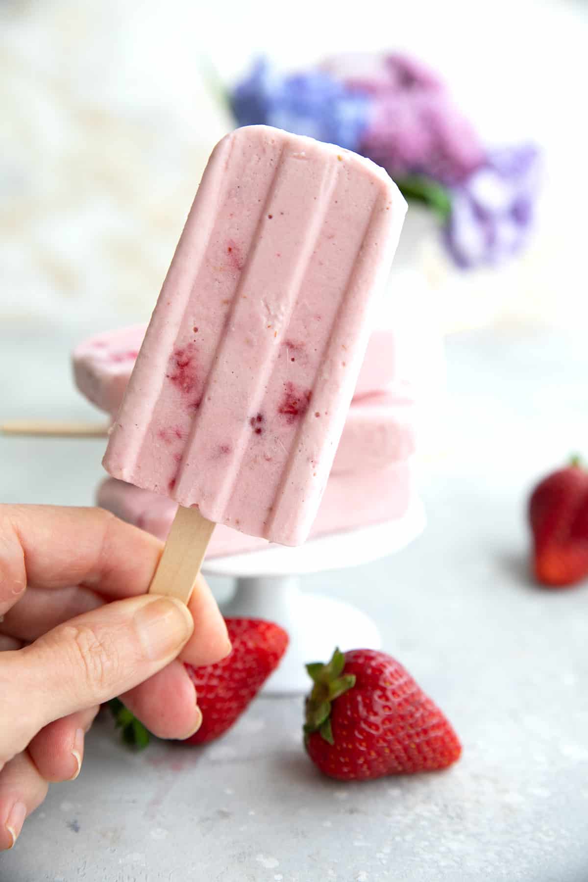A hand holding up a keto strawberry popsicles in front of a table with strawberries and flowers.