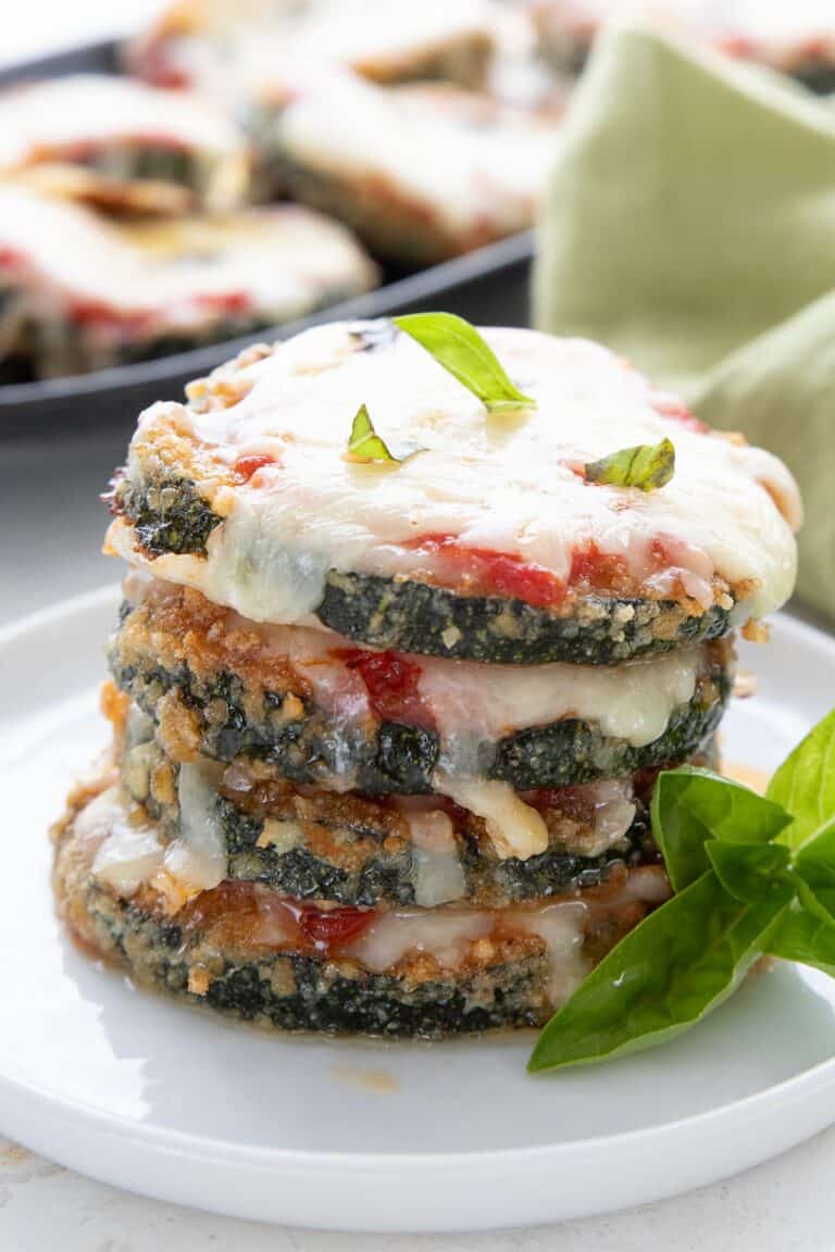 A stack of keto zucchini parmesan on a white plate with a sprig of basil.