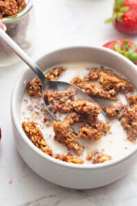 Crunchy keto granola in a white bowl with milk, with a spoonful being lifted out of it.