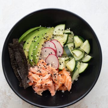 Top down image of salmon, cauliflower rice, cucumber, avocado and radish in a black bowl.