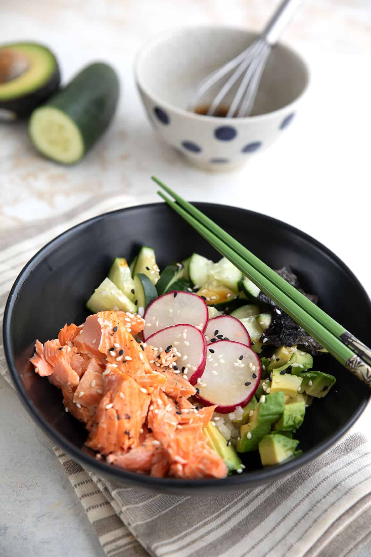 A black bowl filled with keto sushi ingredients, with dressing and vegetables in the background.
