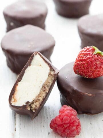 Chocolate covered Keto Cheesecake Bites on a white table with strawberries and raspberries.