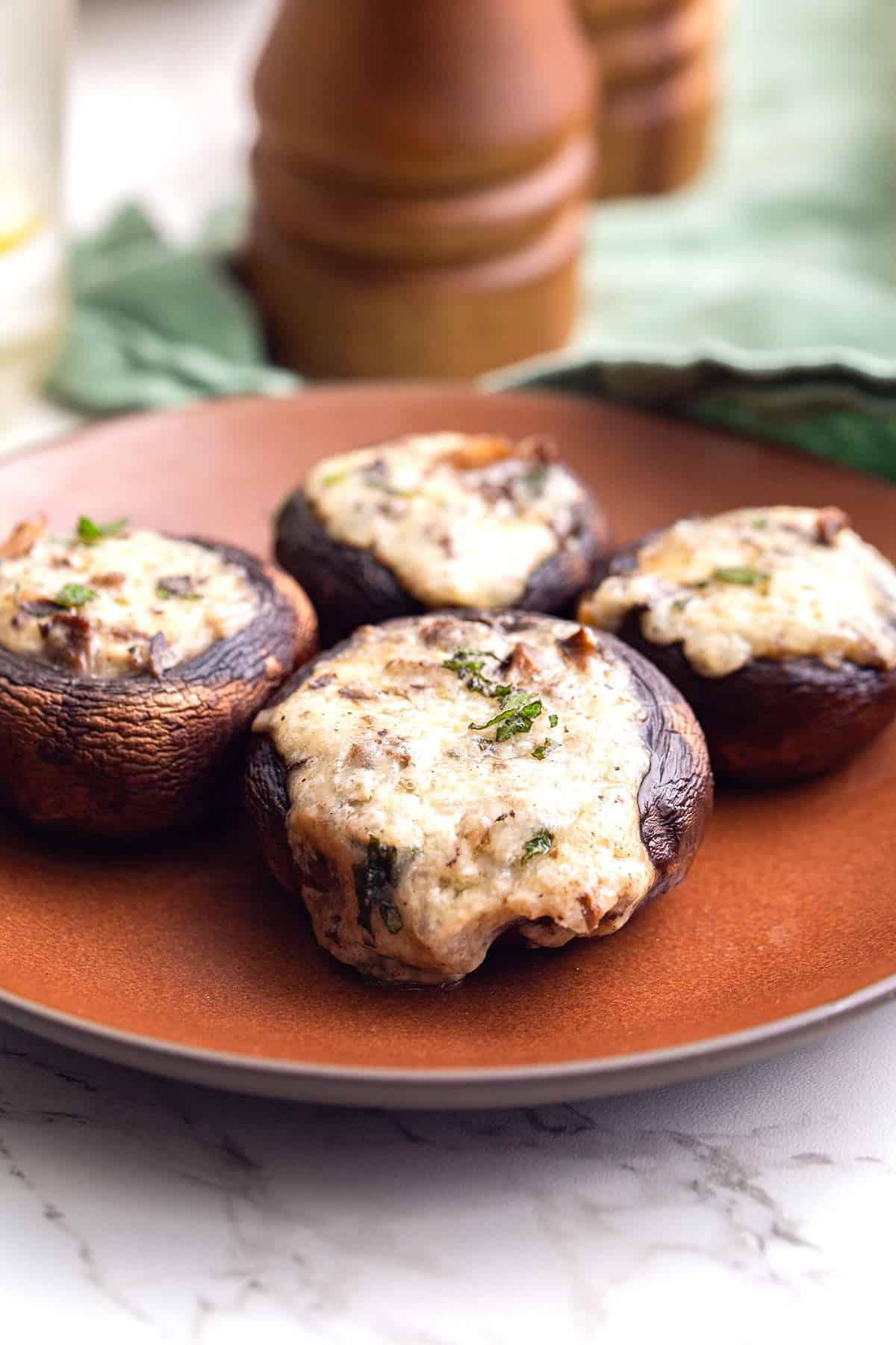 Four Keto Stuffed Mushrooms sit on a brown plate in front of a green napkin.