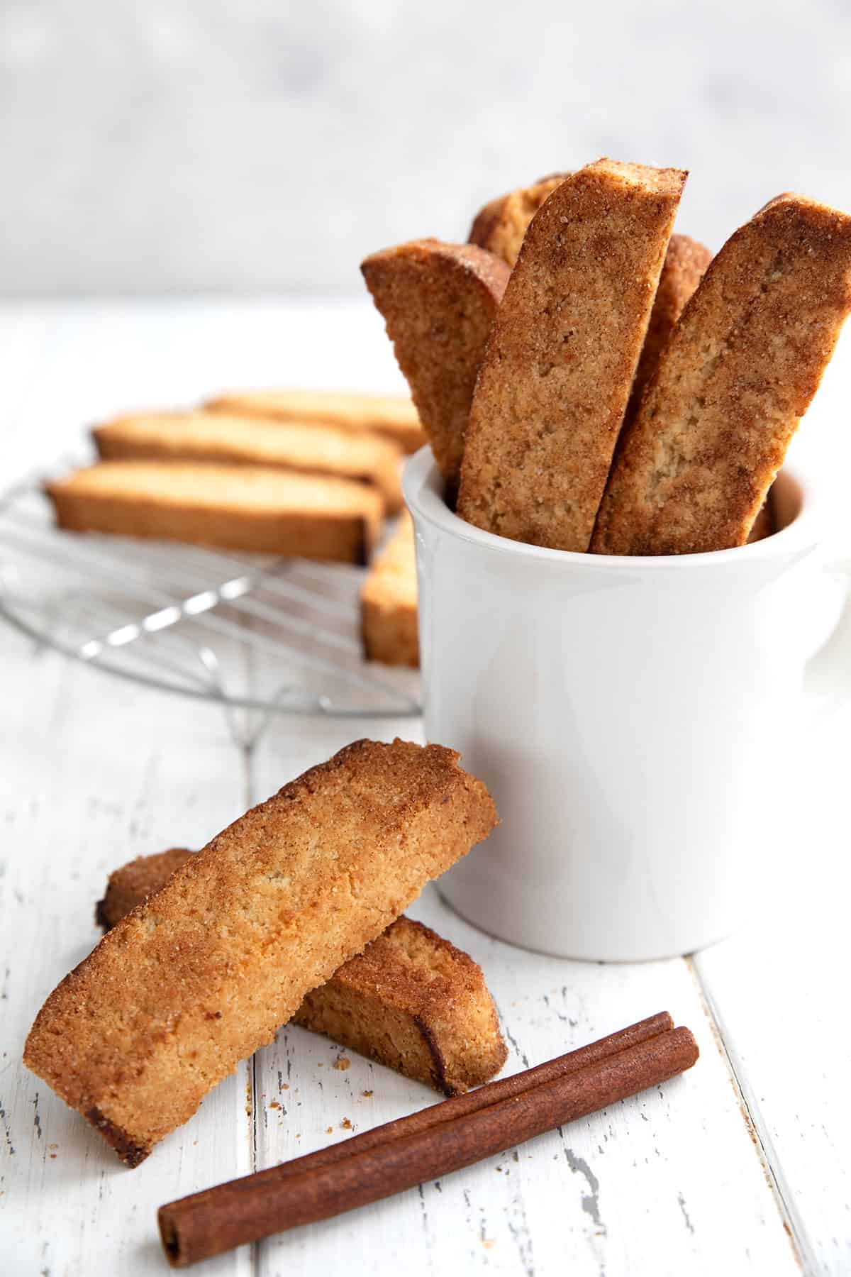 Cinnamon keto biscotti in a white mug on a white table, with two biscotti in front.