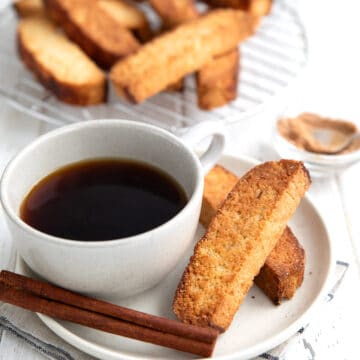 Two keto biscotti on a white plate with a cup of coffee and a cinnamon stick.