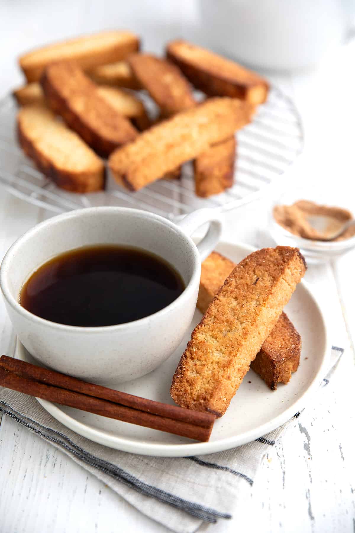 Two keto biscotti on a white plate with a cup of coffee and a cinnamon stick.