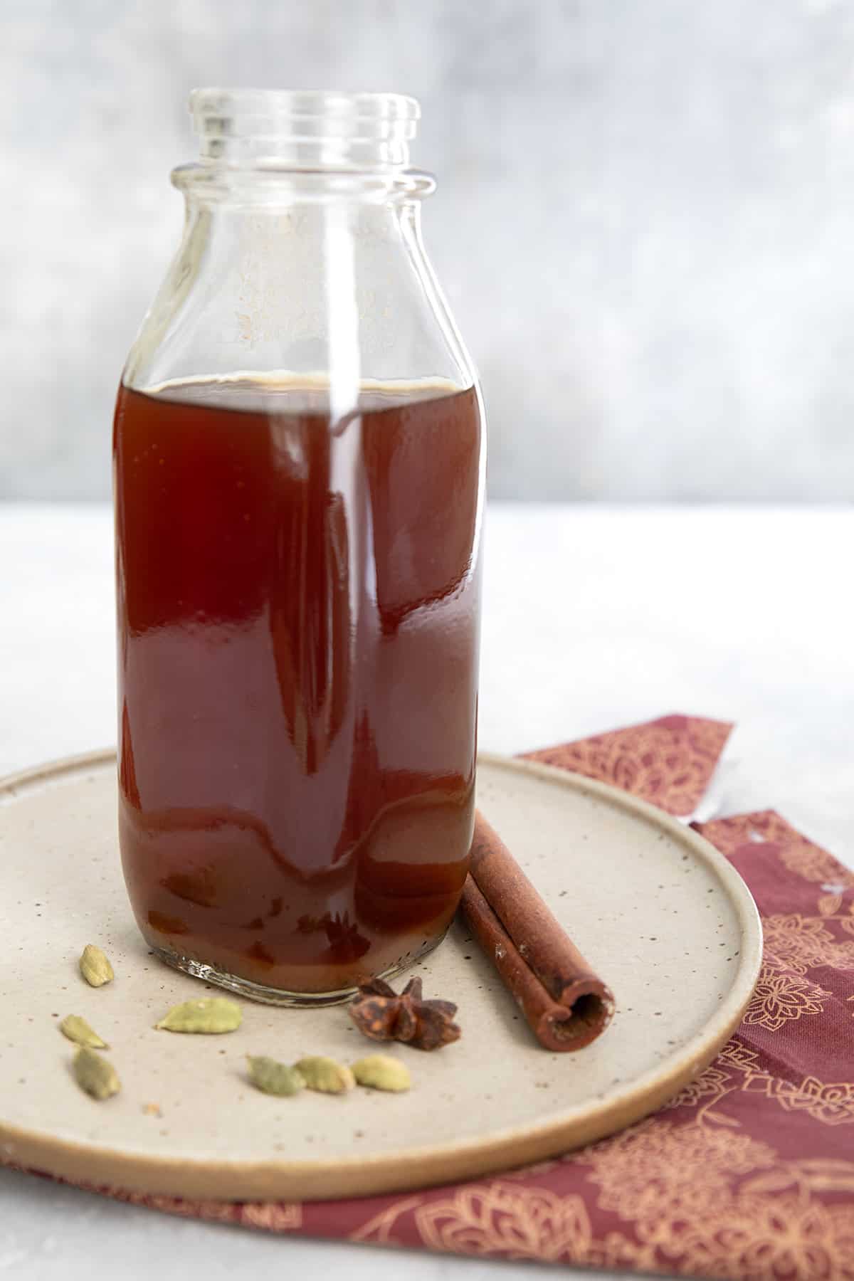 A bottle of keto chai concentrate on a rustic plate over a white wooden table, with cardamom pods and cinnamon sticks.