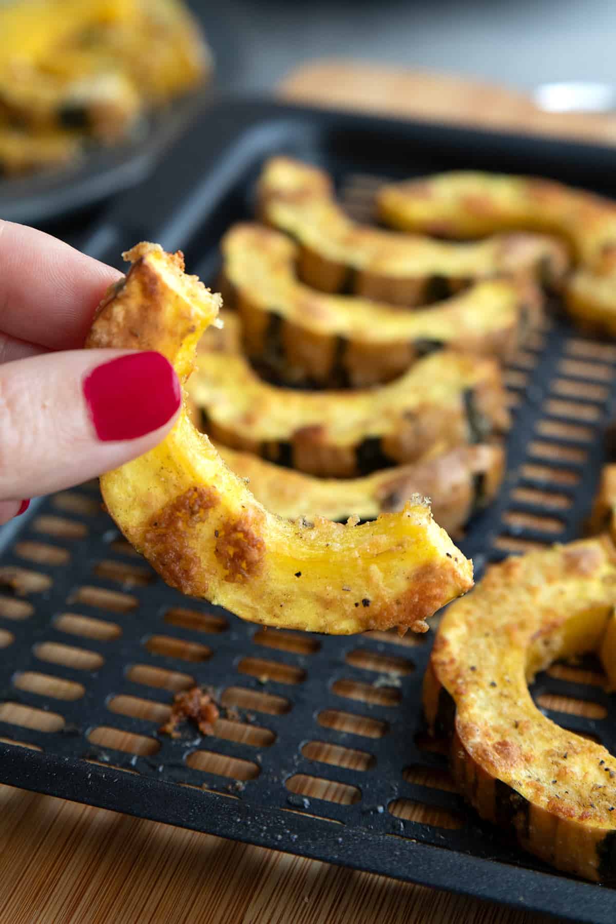 A hand holding up a piece of Roasted Delicata Squash from an air fryer tray.