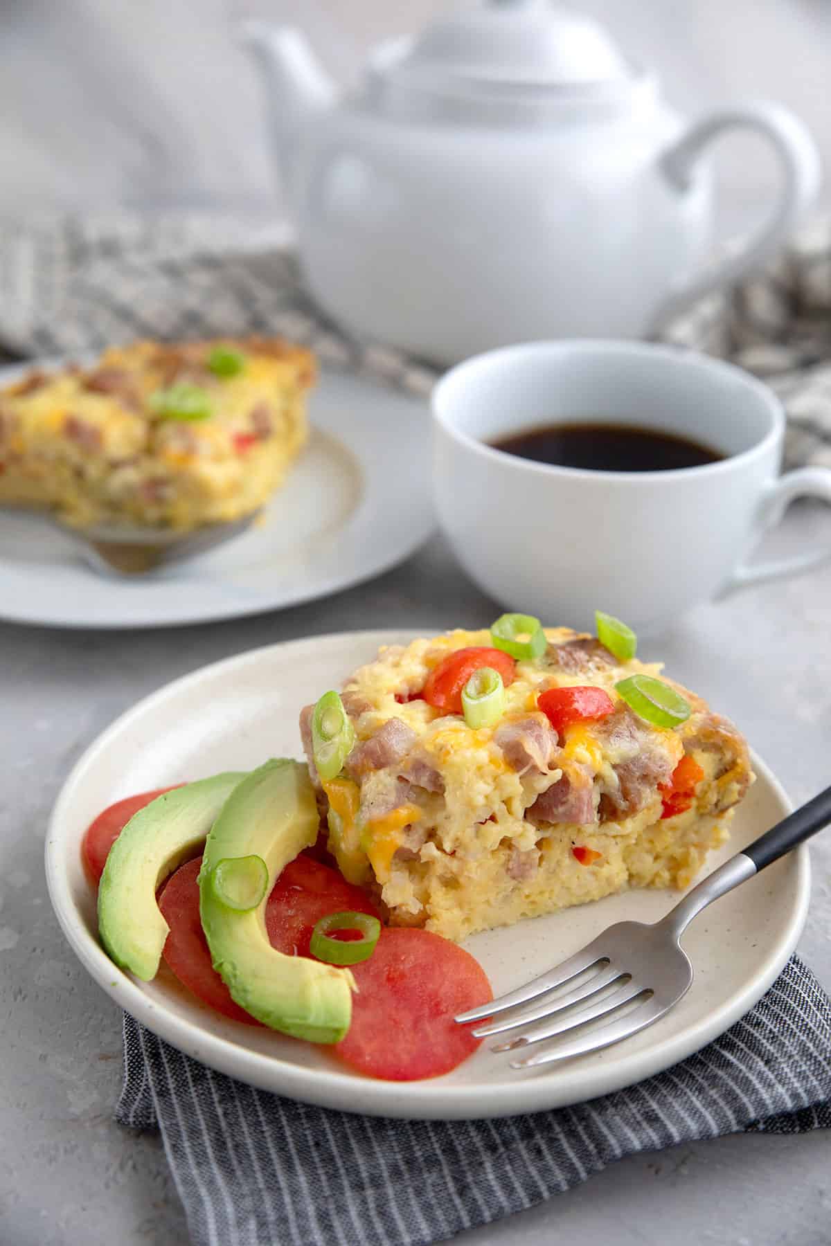 Two slices of Slow Cooker Breakfast Casserole on white plates, with a cup of coffee and a coffee pot in the background.