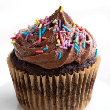 A single chocolate keto cupcake on a white cupcake stand with sugar free chocolate frosting and sprinkles on top.