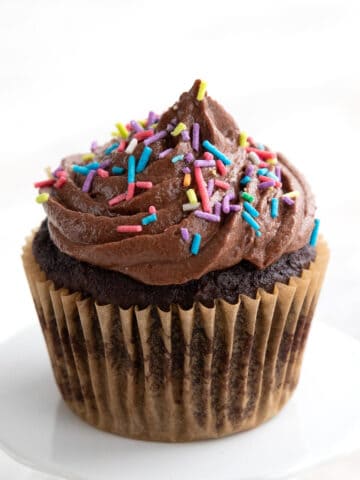 A single chocolate keto cupcake on a white cupcake stand with sugar free chocolate frosting and sprinkles on top.