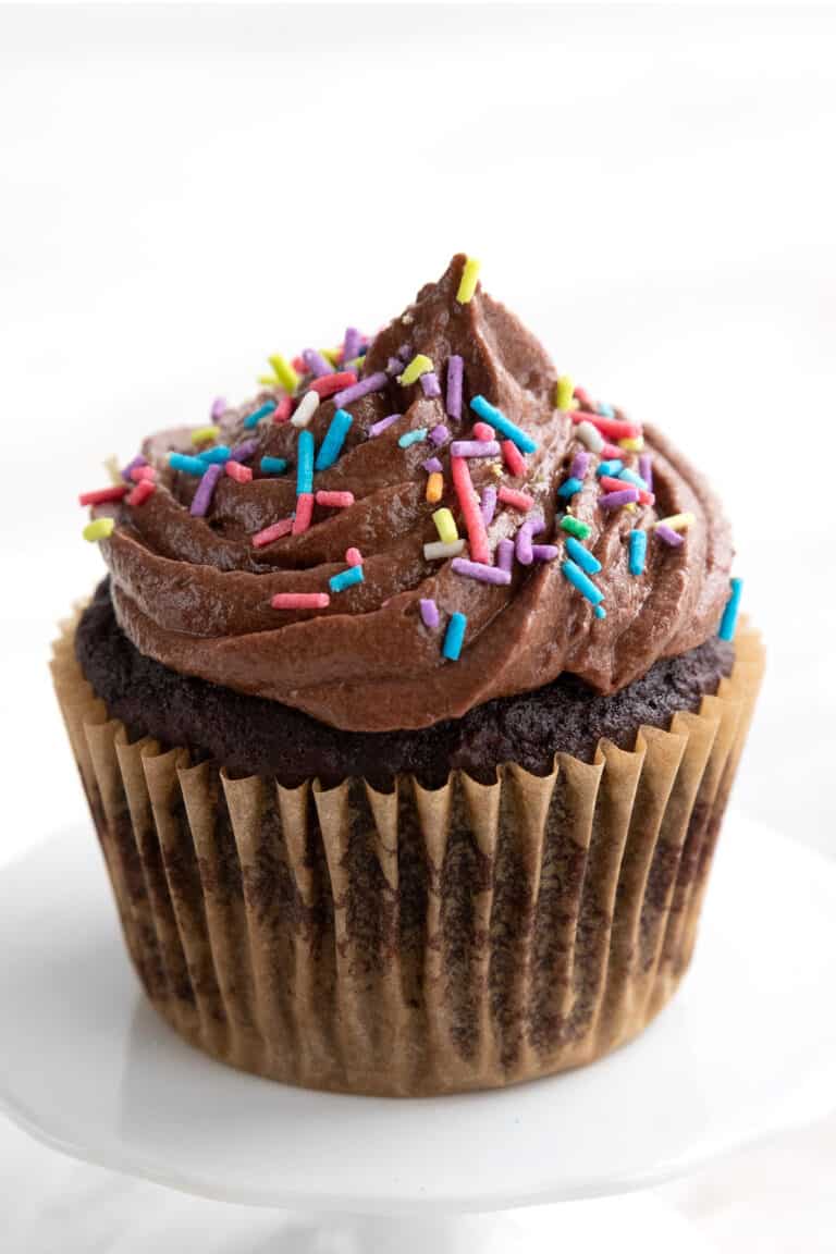 A single chocolate keto cupcake on a white cupcake stand with sugar free chocolate frosting and sprinkles on top.