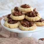 A stack of Keto Pecan Pie Cookies on a white cake stand with pecans strewn around.