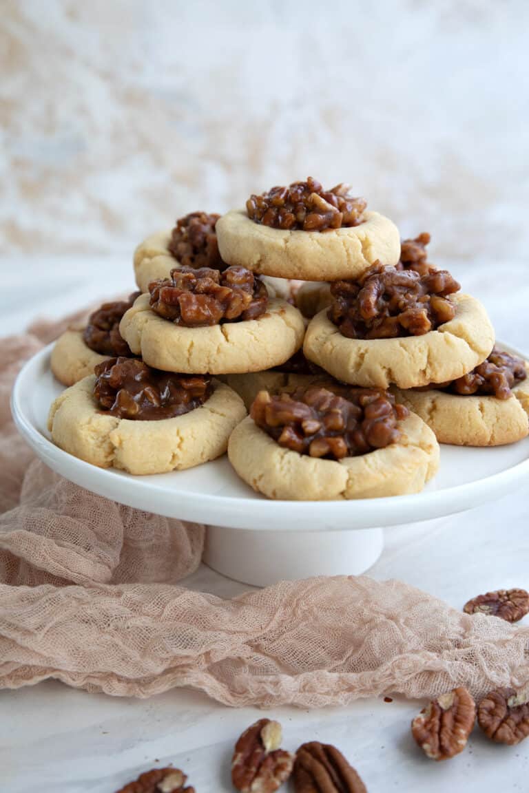 A stack of Keto Pecan Pie Cookies on a white cake stand with pecans strewn around.