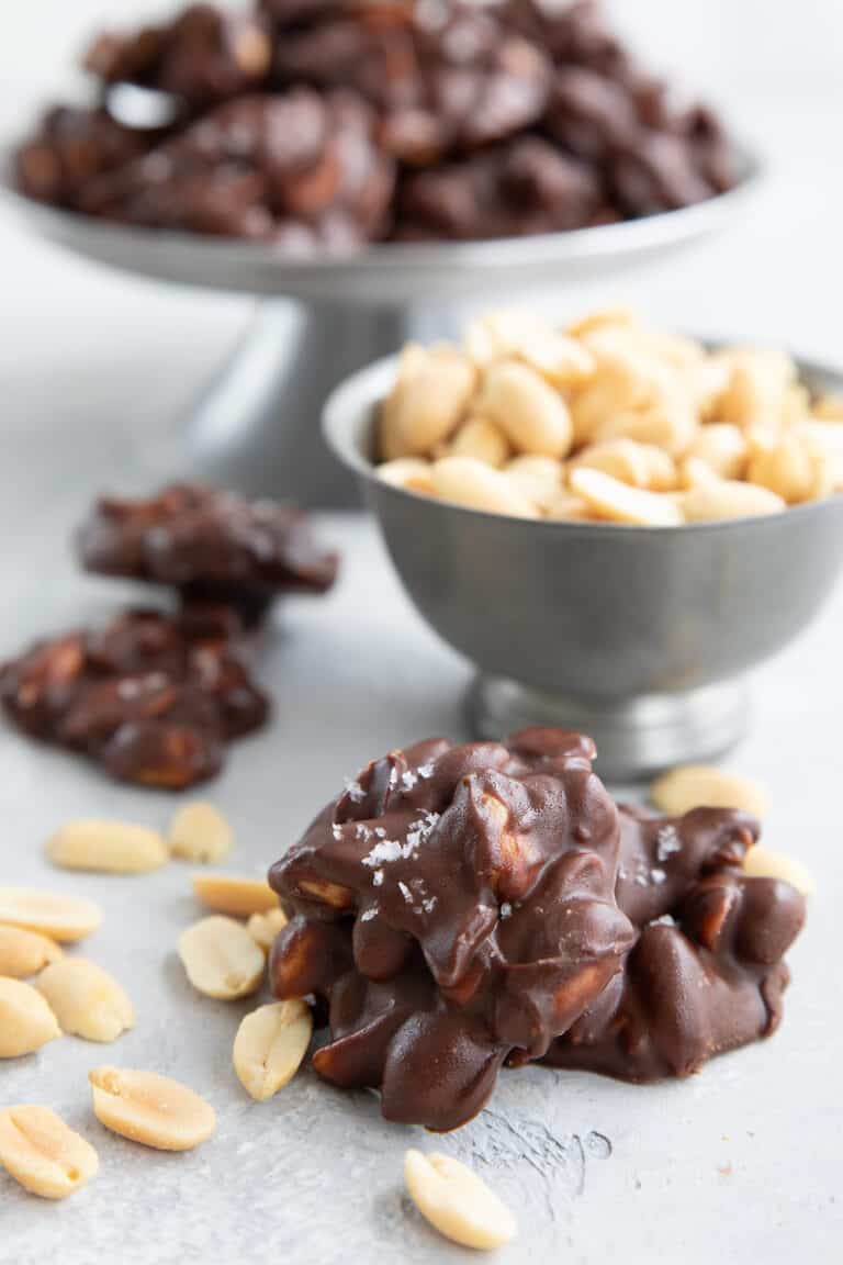 Two Keto Peanut Clusters with a bowl of peanuts more clusters in the background.