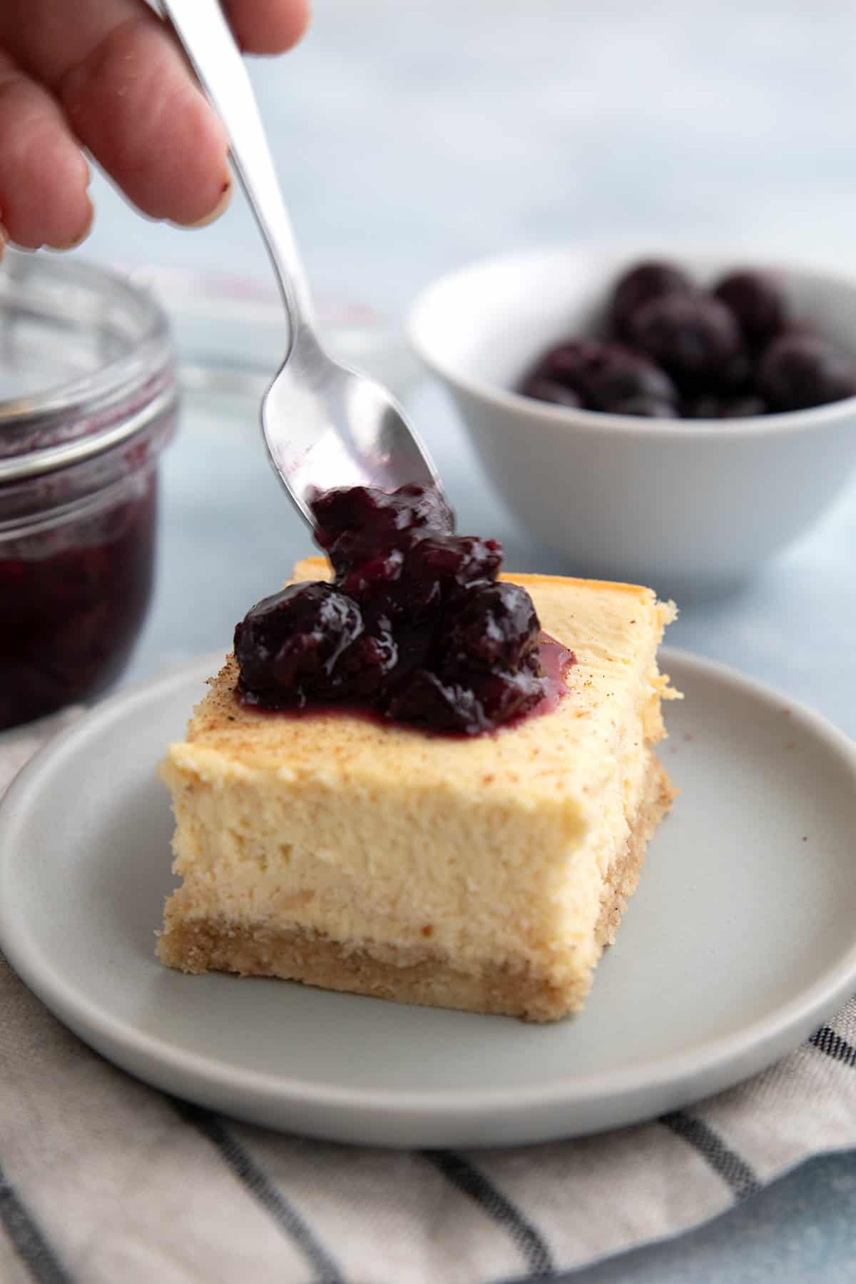A spoon drizzling keto blueberry syrup over keto cheesecake.