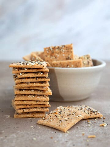 A stack of crispy keto crackers on a brown table with a bowl of crackers in behind it.