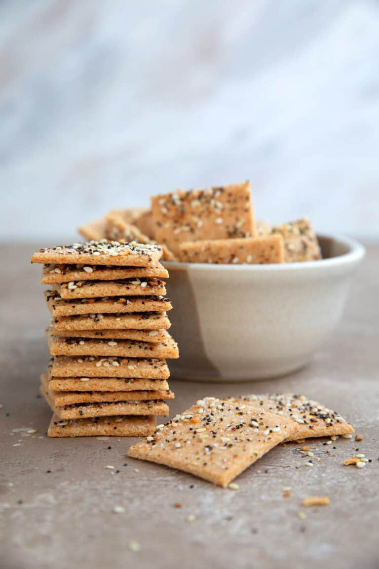 A stack of crispy keto crackers on a brown table with a bowl of crackers in behind it.