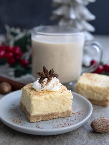 A keto eggnog cheesecake bar with whipped cream and star anise on top, and a glass of eggnog in the background.