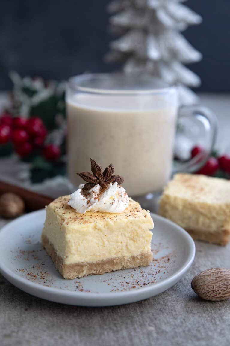 A keto eggnog cheesecake bar with whipped cream and star anise on top, and a glass of eggnog in the background.