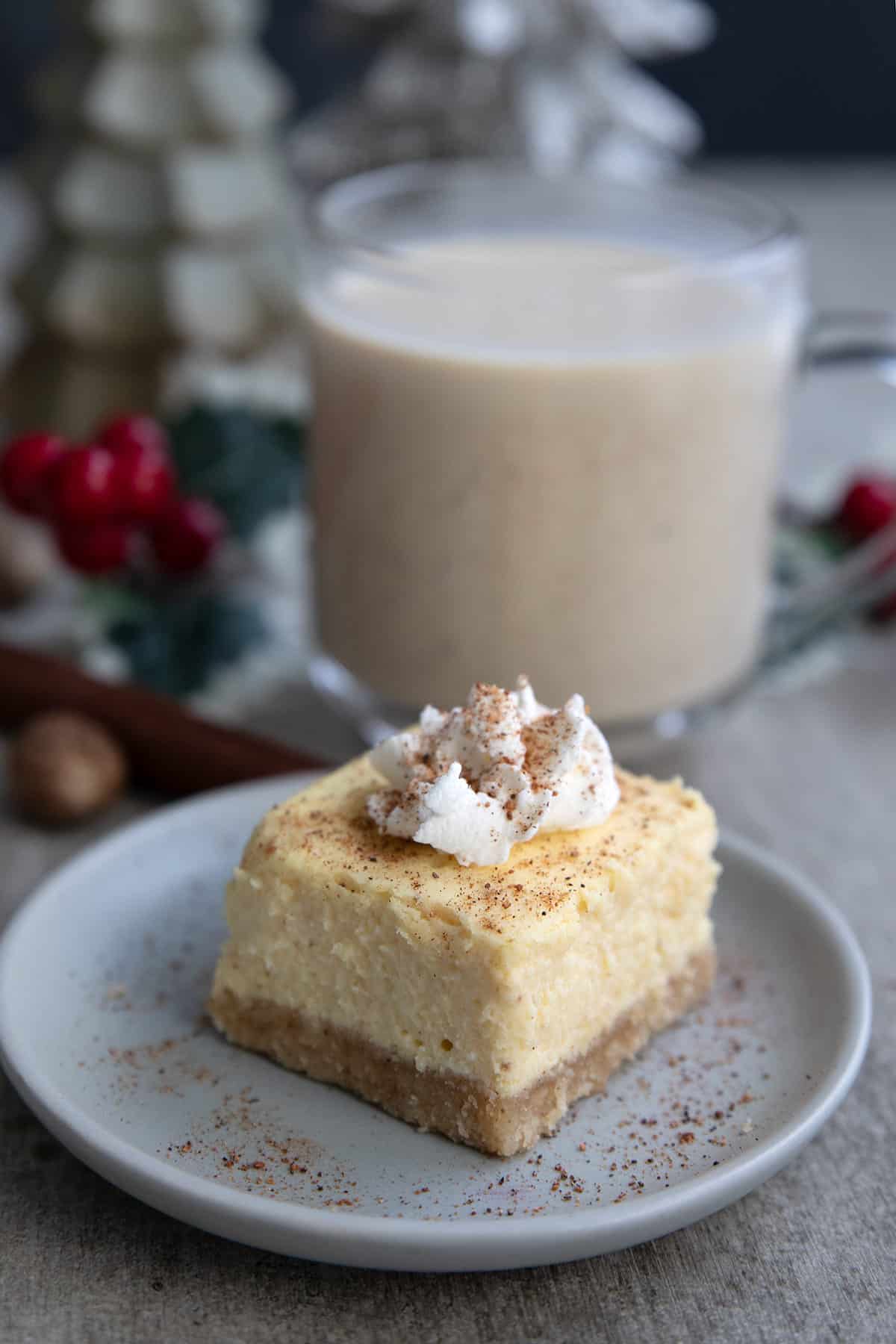 A keto eggnog cheesecake bar sits on a small dessert plate, in front of a cup of eggnog.