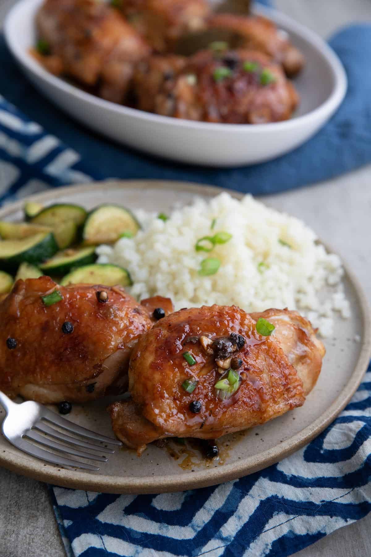 Chicken Adobo on a plate with cauliflower rice and sautéed zucchini, over a blue patterned napkin.