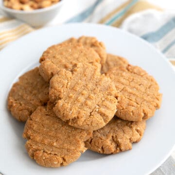 Keto Peanut Butter Cookies piled on a white plate with a small bowl of peanuts in the background.