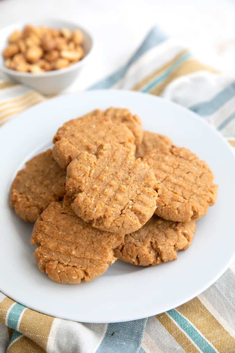 Keto Peanut Butter Cookies piled on a white plate with a small bowl of peanuts in the background.