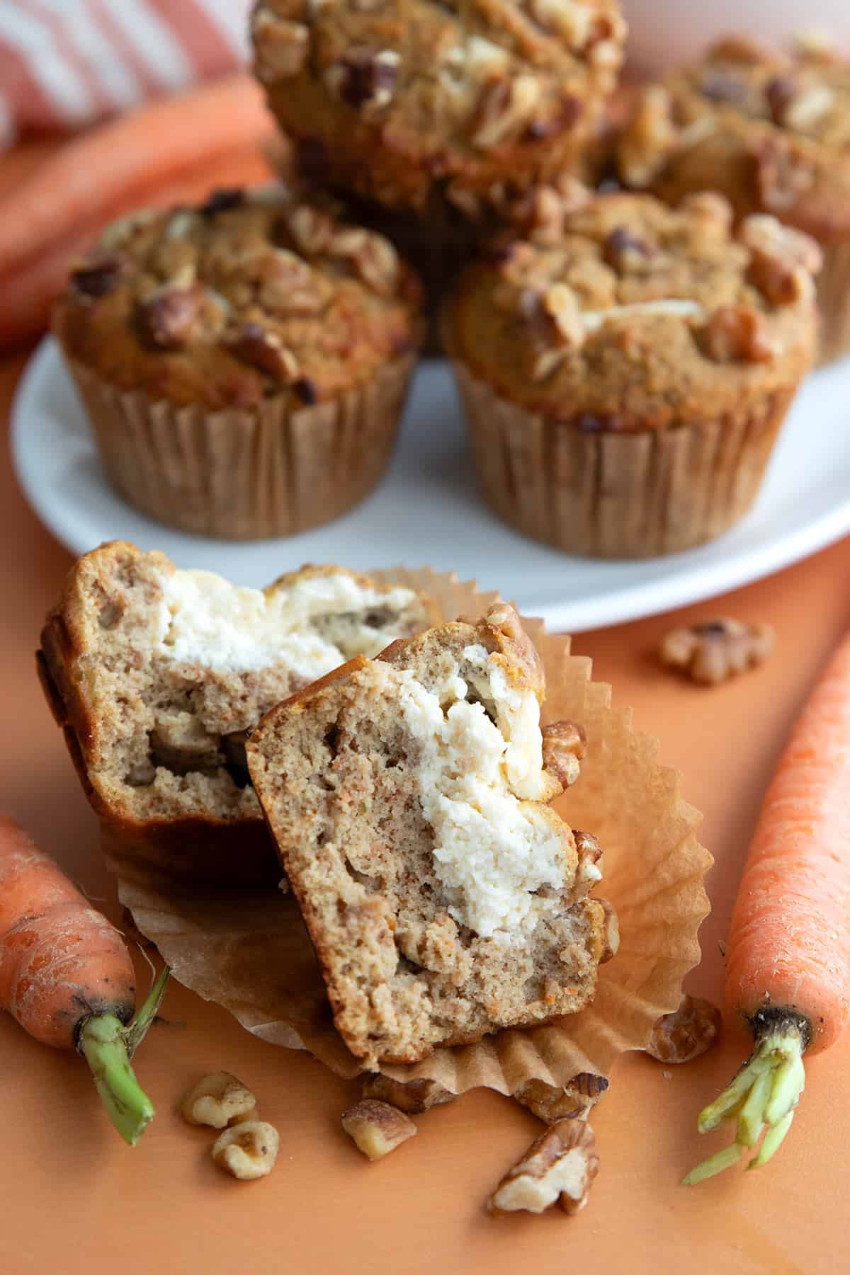 A keto carrot cake muffin split open in front of a plate of more muffins, with carrots and walnuts strewn around.