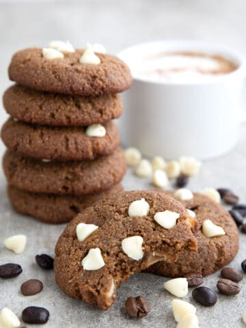 Two Keto Cappuccino Cookies in front of a stack of more cookies, with white chocolate chips and espresso beans strewn around.