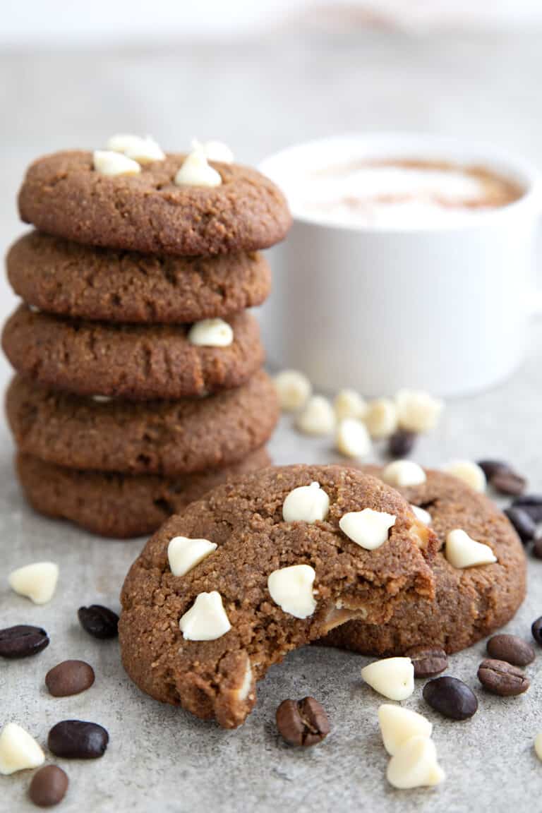 Two Keto Cappuccino Cookies in front of a stack of more cookies, with white chocolate chips and espresso beans strewn around.