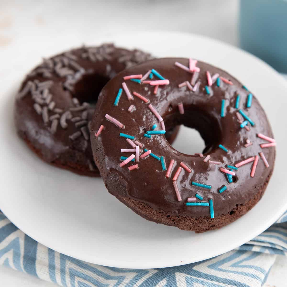 Two keto chocolate donuts with chocolate glaze and sprinkles on a white plate over a blue patterned napkin.
